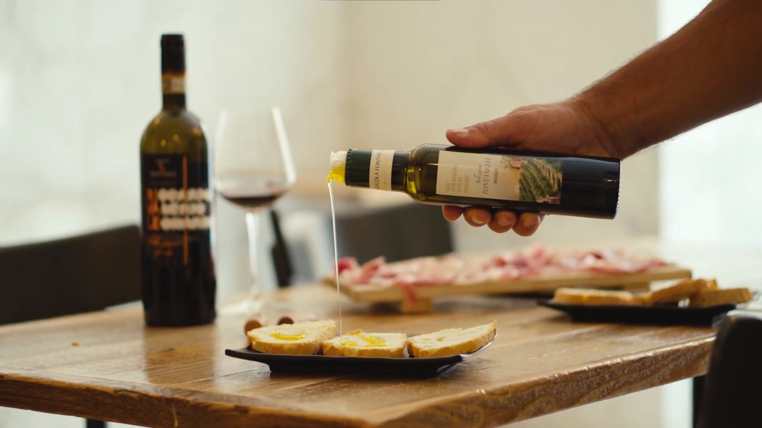 Olive oil being poured onto toasted bread with a bottle and glass of wine in the background